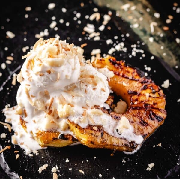 Grilled pineapple slices topped with ice cream and crumbled coconut and macadamia nuts on a black plate.